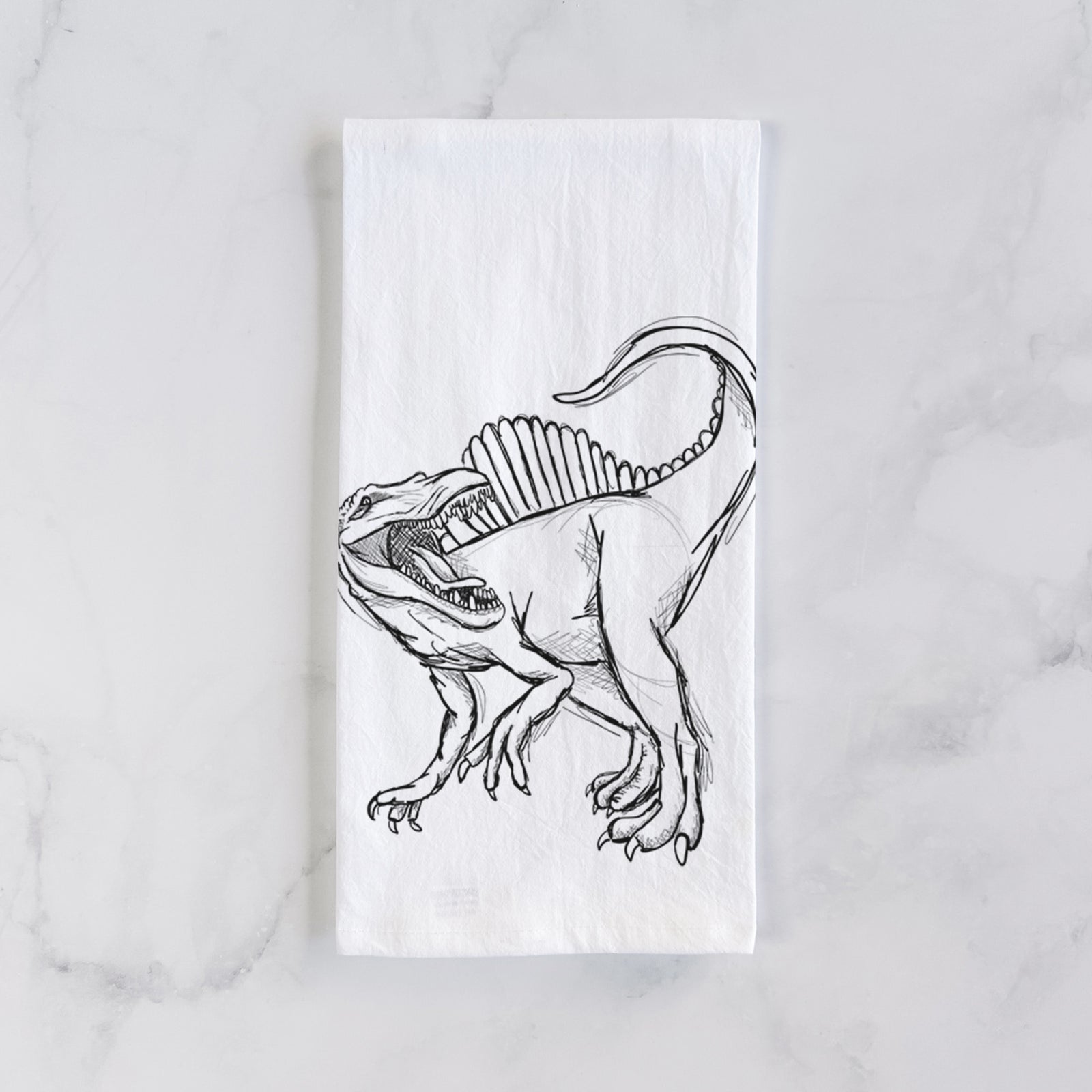 White cotton tea towel printed with a black ink illustration of a Spinosaurus with its tall sail-shaped back, on a marble counter beside a round wooden cutting board and greenery.