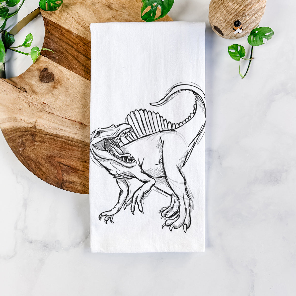 White cotton tea towel printed with a black ink illustration of a Spinosaurus with its tall sail-shaped back, on a marble counter beside a round wooden cutting board and greenery.