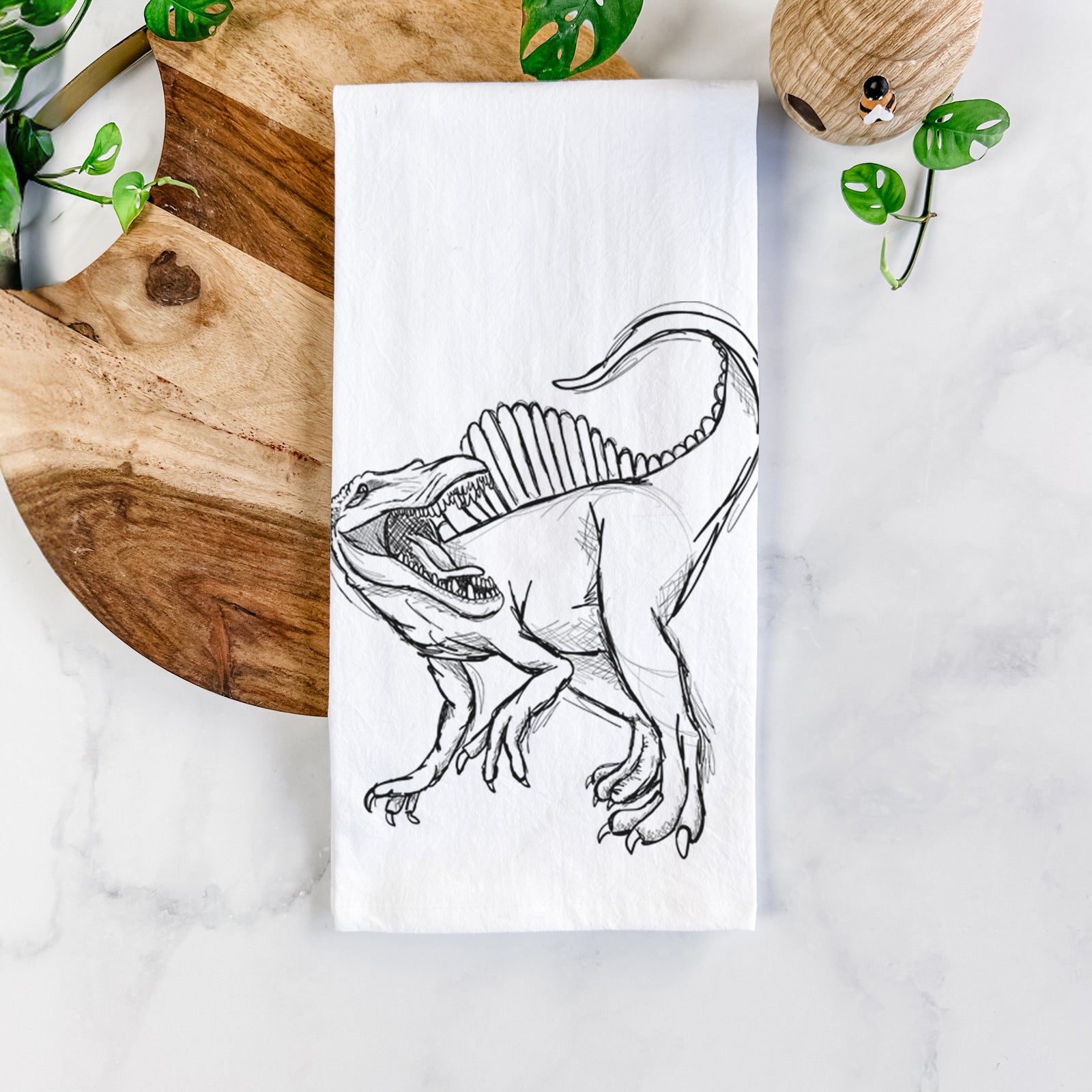 White cotton tea towel printed with a black ink illustration of a Spinosaurus with its tall sail-shaped back, on a marble counter beside a round wooden cutting board and greenery.