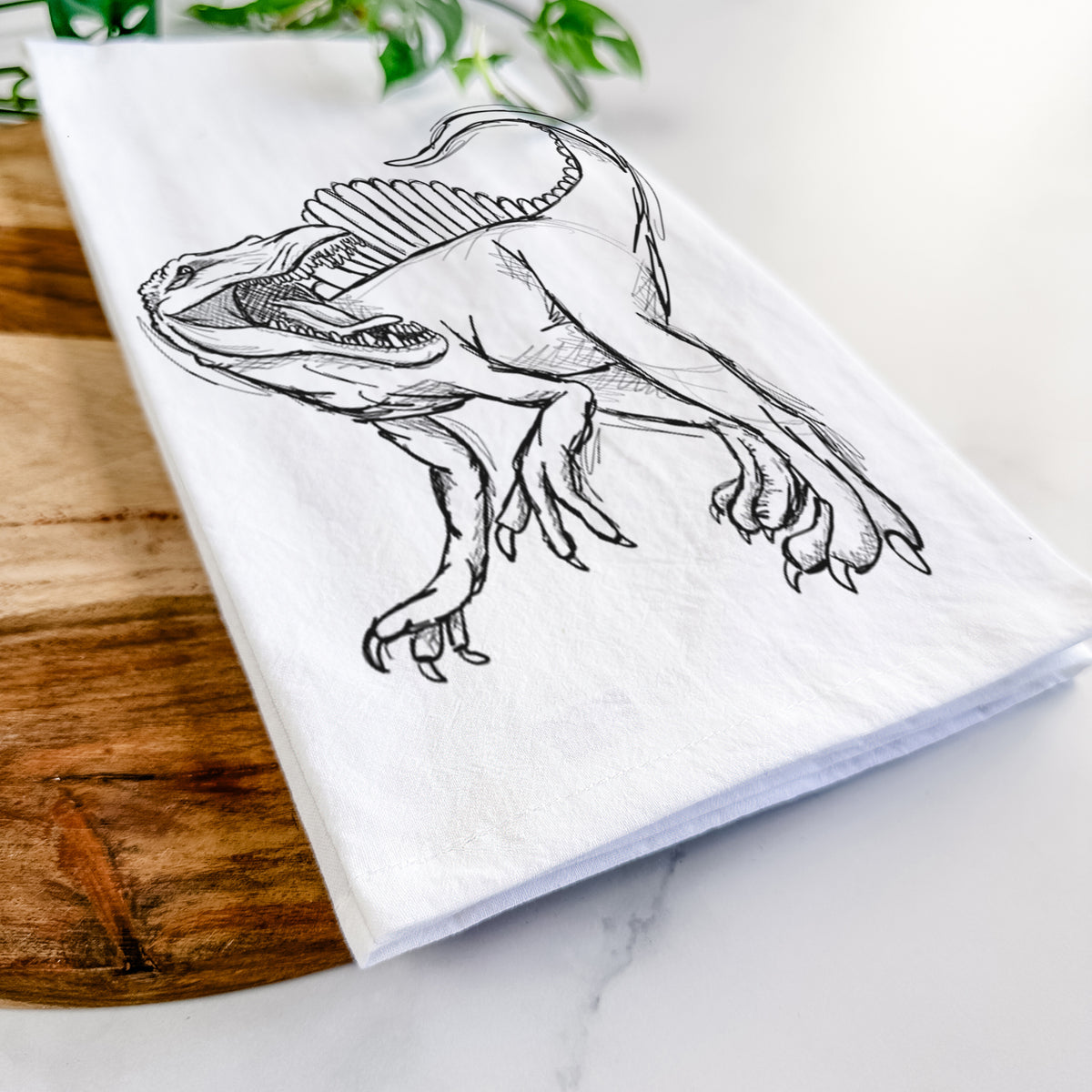 Close-up of a folded white tea towel on a wooden board, showing a black ink illustration of a Spinosaurus with its tall sail-shaped back.