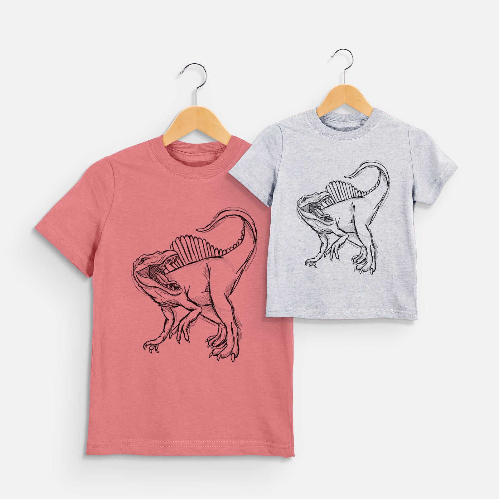 Two youth t-shirts in different colors on hangers, printed with a black ink illustration of a Spinosaurus with its tall sail-shaped back.