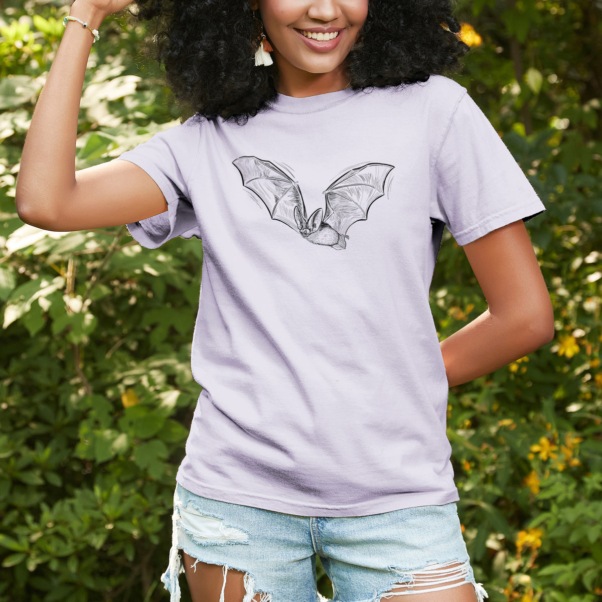 Woman wearing a Comfort Colors t-shirt printed with a black ink illustration of a spotted bat in flight with large ears, outdoors.