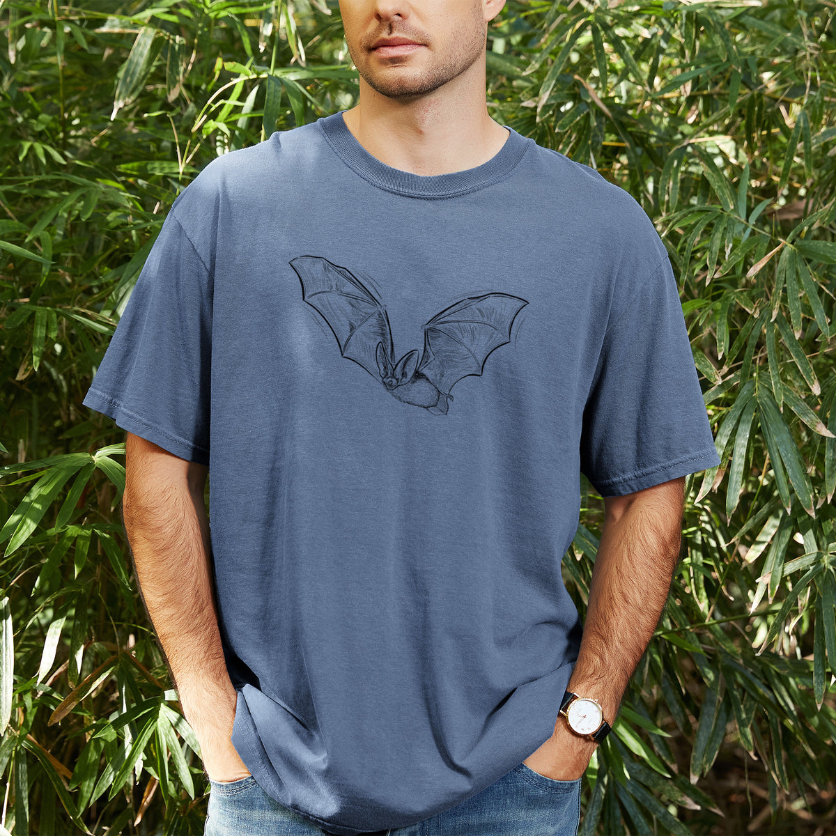 Man wearing a Comfort Colors t-shirt printed with a black ink illustration of a spotted bat in flight with large ears, outdoors.