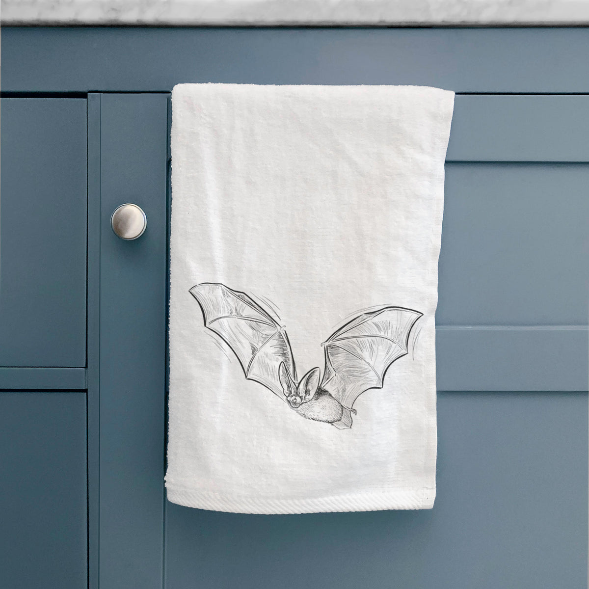 White hand towel printed with a black ink illustration of a spotted bat in flight with large ears, hanging on a cabinet door.