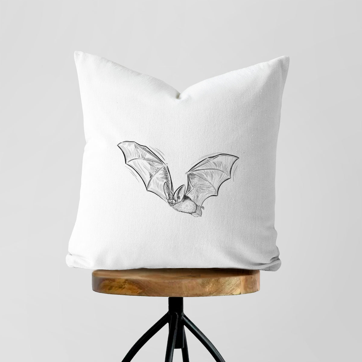 White pillow cover printed with a black ink illustration of a spotted bat in flight with large ears, on a wooden stool.