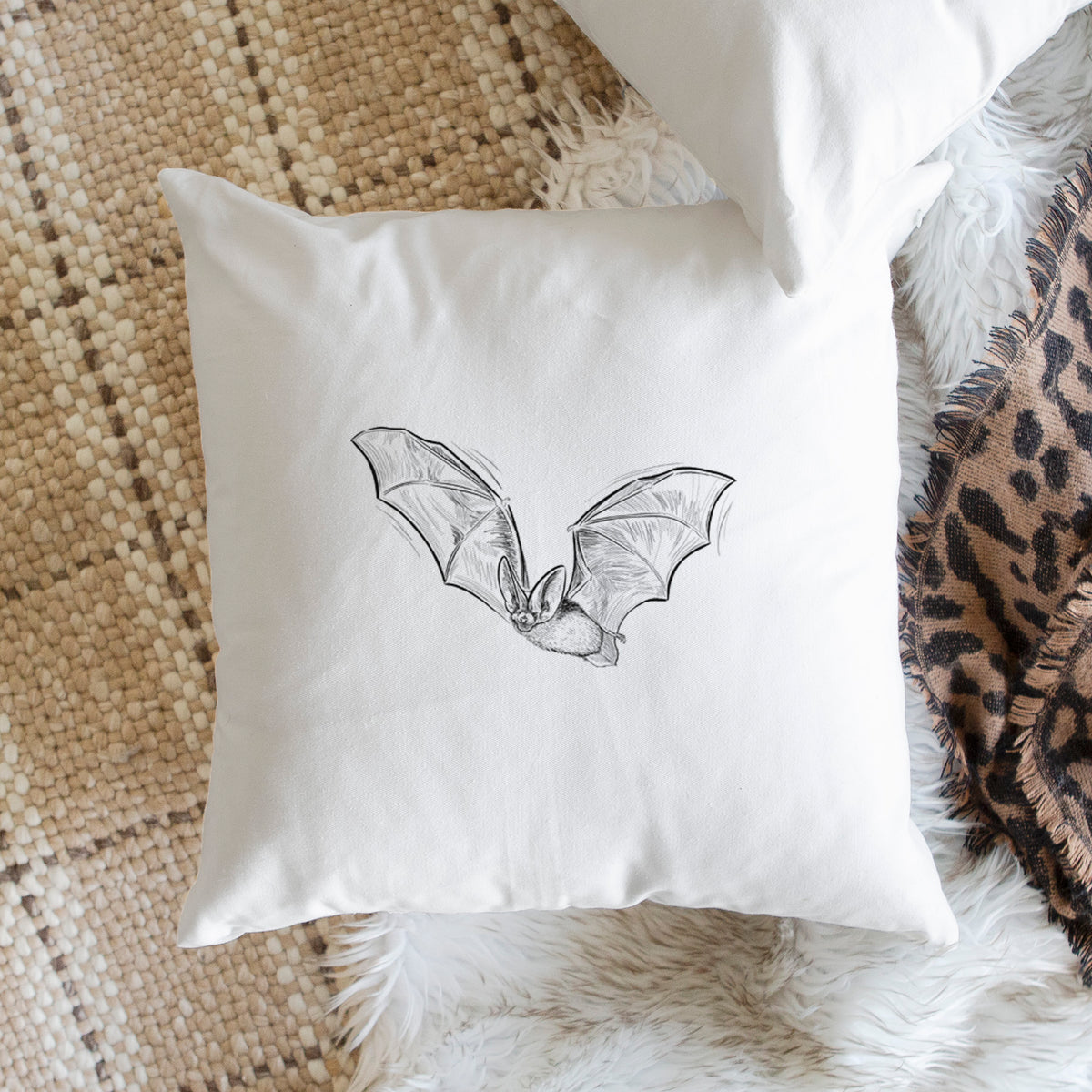 White pillow cover printed with a black ink illustration of a spotted bat in flight with large ears, on a woven chair.