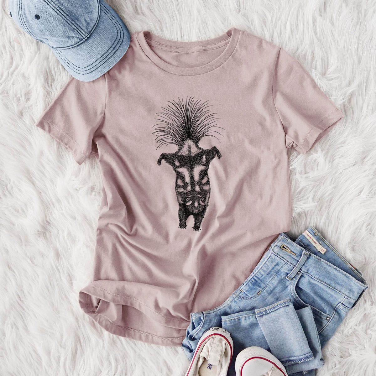 Women's relaxed fit cotton t-shirt printed with a black ink illustration of a spotted skunk doing a handstand with its tail fanned, styled flat on a white faux fur rug with jeans.