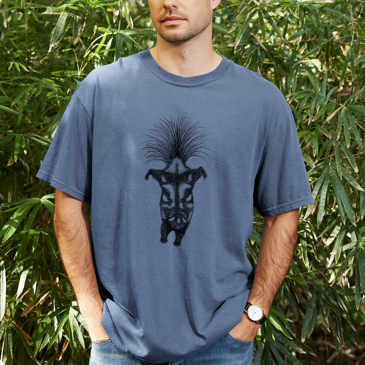 Man wearing a Comfort Colors t-shirt printed with a black ink illustration of a spotted skunk doing a handstand with its tail fanned, outdoors.
