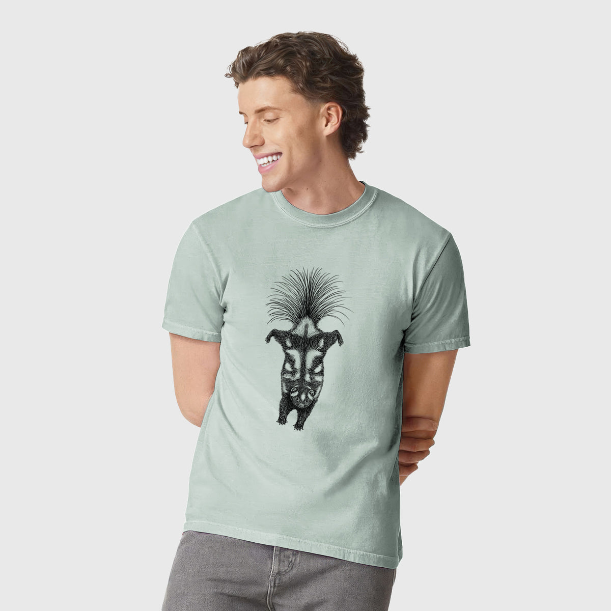 Man wearing a Comfort Colors t-shirt printed with a black ink illustration of a spotted skunk doing a handstand with its tail fanned.