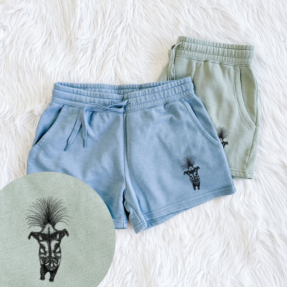Two pairs of Cali Wave shorts in different colors printed with a black ink illustration of a spotted skunk doing a handstand with its tail fanned, with a close-up of the print.