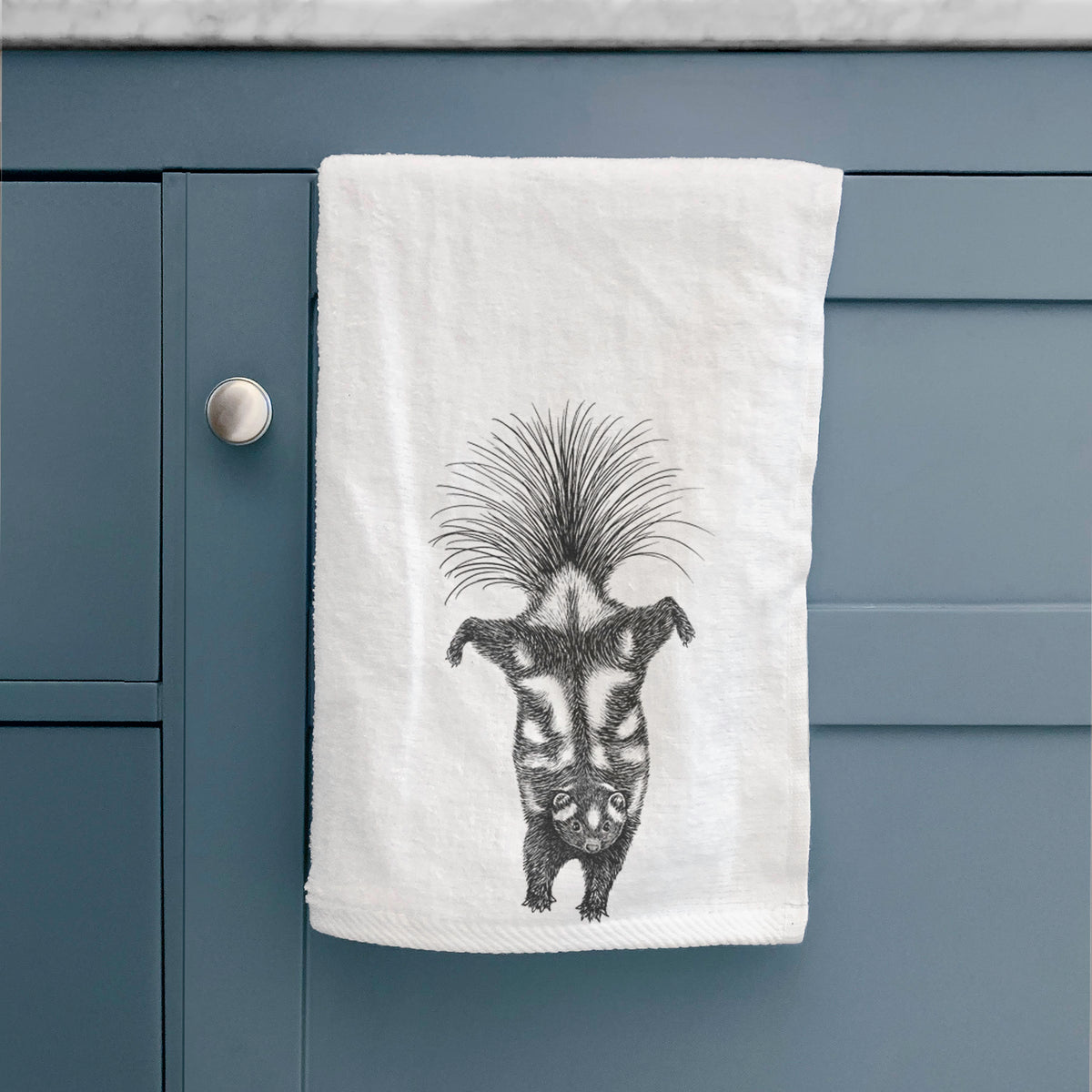 White hand towel printed with a black ink illustration of a spotted skunk doing a handstand with its tail fanned, hanging on a cabinet door.