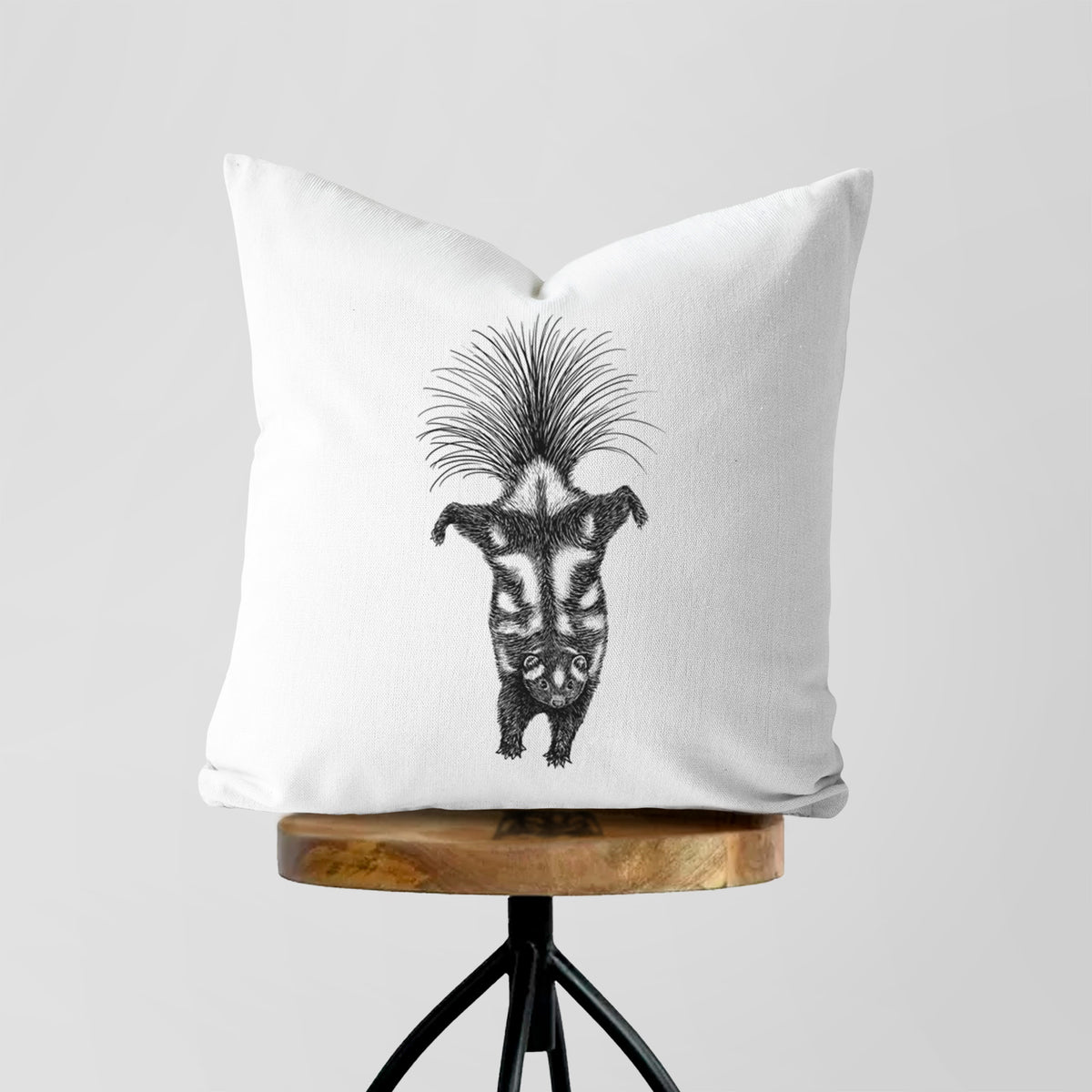 White pillow cover printed with a black ink illustration of a spotted skunk doing a handstand with its tail fanned, on a wooden stool.