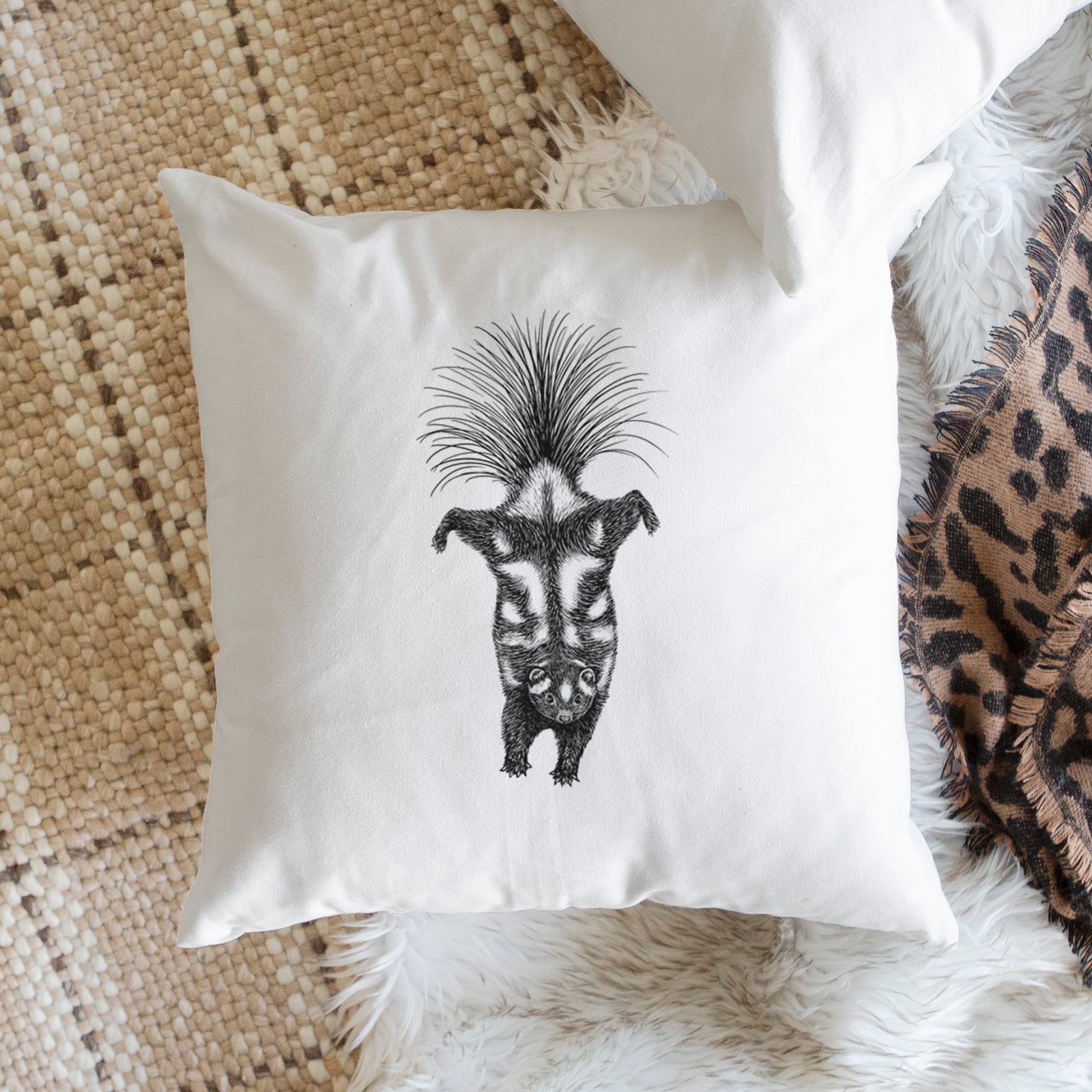 White pillow cover printed with a black ink illustration of a spotted skunk doing a handstand with its tail fanned, on a bench beside plants.