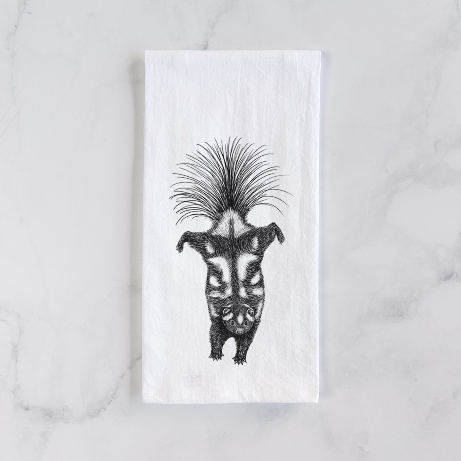 White cotton tea towel printed with a black ink illustration of a spotted skunk doing a handstand with its tail fanned, on a marble counter beside a round wooden cutting board and greenery.