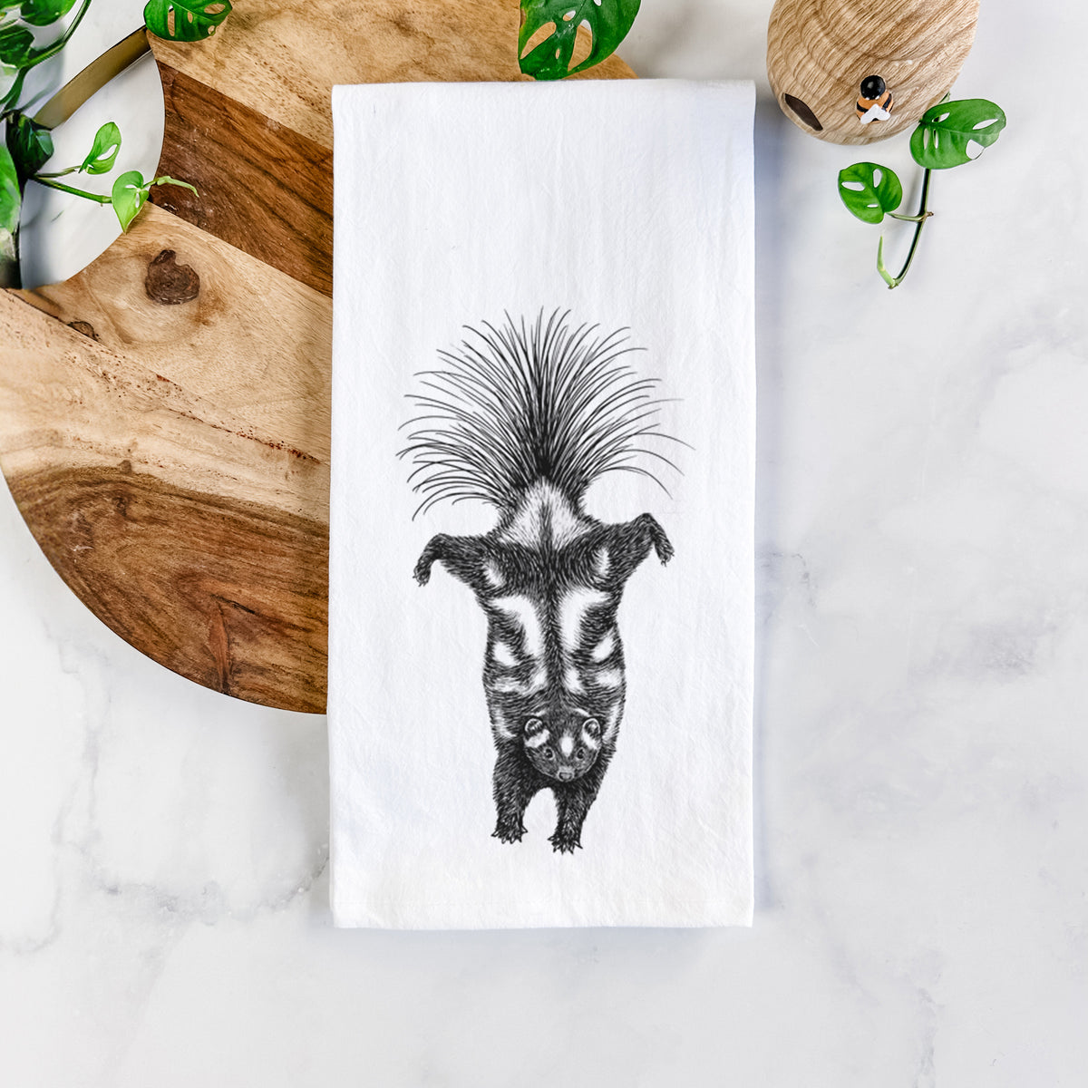 White cotton tea towel printed with a black ink illustration of a spotted skunk doing a handstand with its tail fanned, on a marble counter beside a round wooden cutting board and greenery.