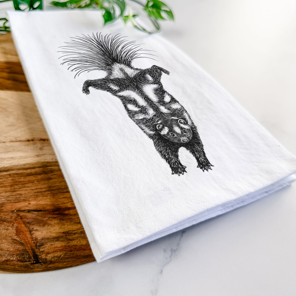 Close-up of a folded white tea towel on a wooden board, showing a black ink illustration of a spotted skunk doing a handstand with its tail fanned.
