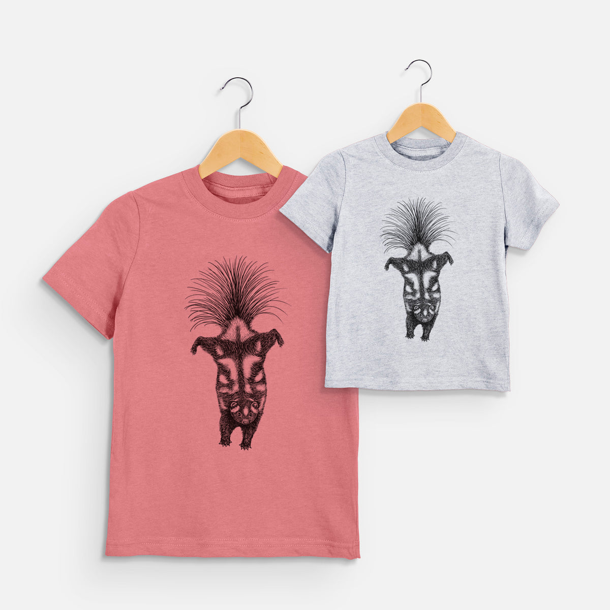 Two youth t-shirts in different colors on hangers, printed with a black ink illustration of a spotted skunk doing a handstand with its tail fanned.