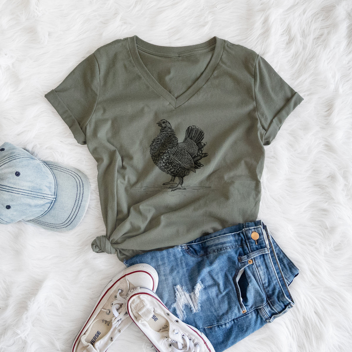 Military green women's relaxed fit cotton V-neck t-shirt printed with a detailed black ink illustration of a spruce grouse standing with its tail fanned, styled flat on a white faux fur rug with a cap, jeans and sneakers.