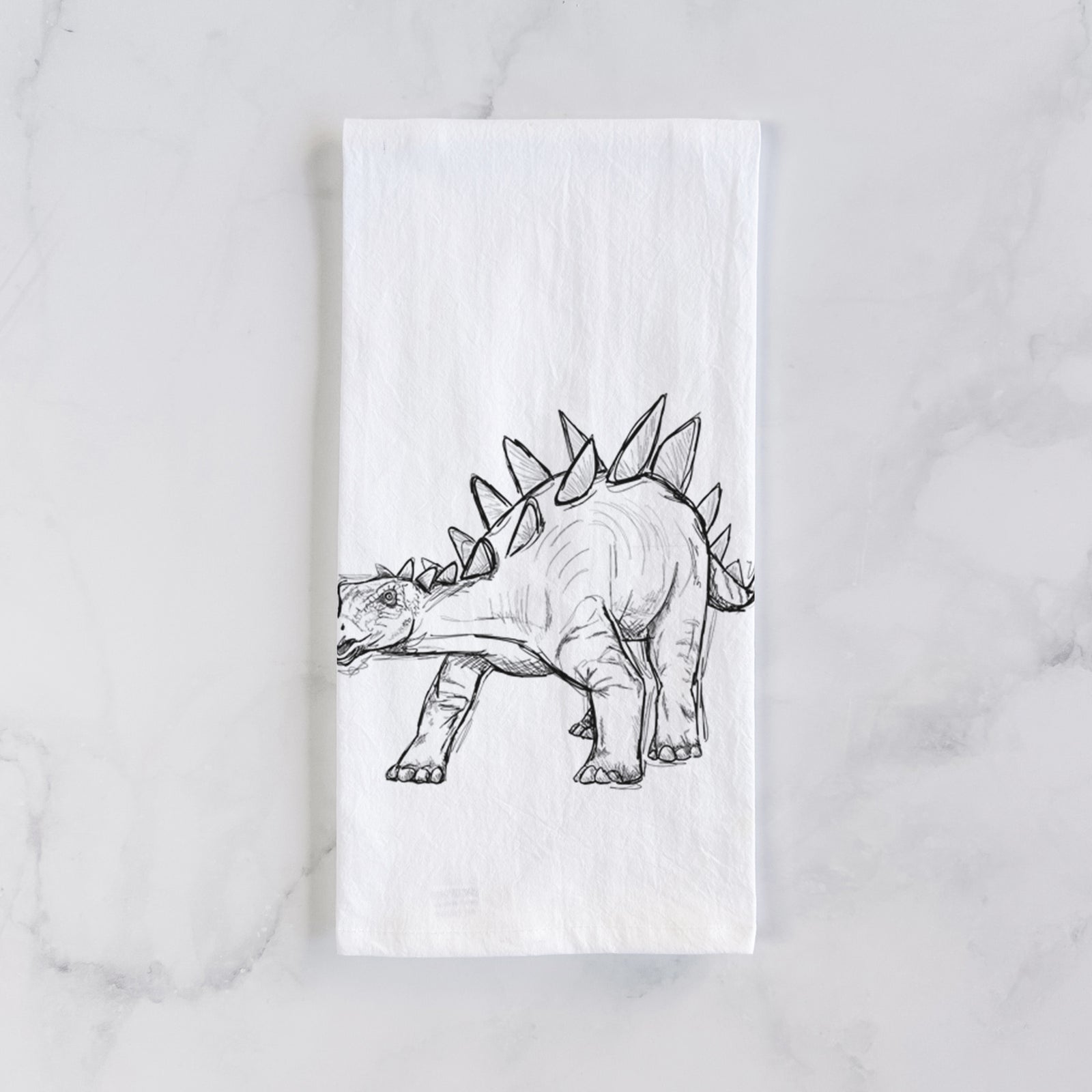White cotton tea towel printed with a black ink illustration of a Stegosaurus with plates along its back, on a marble counter beside a round wooden cutting board and greenery.