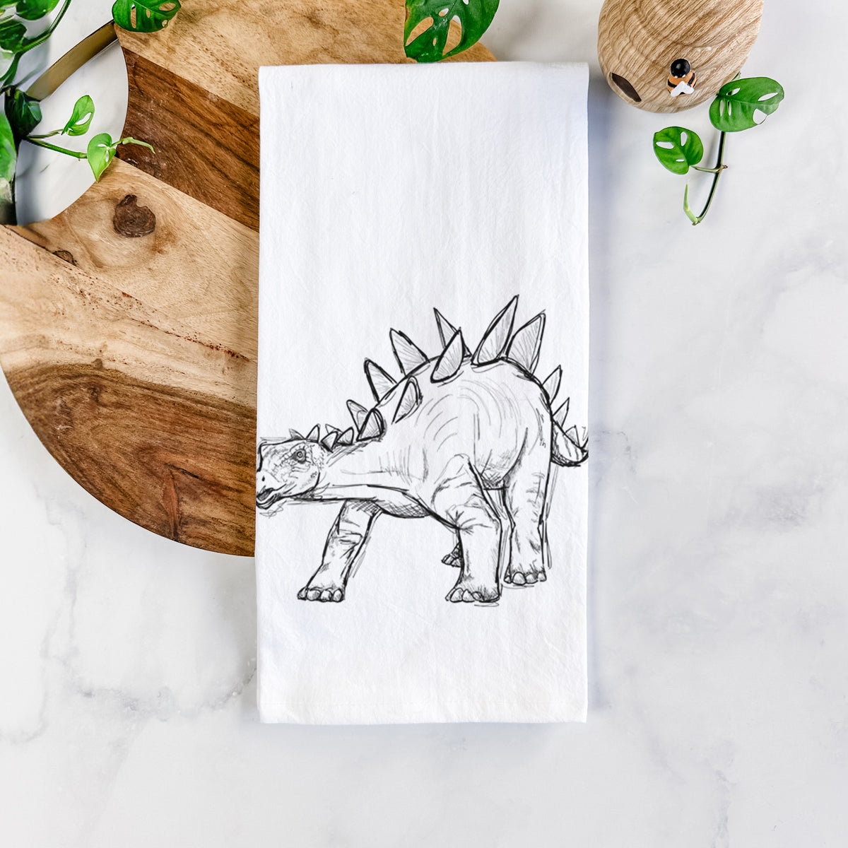 White cotton tea towel printed with a black ink illustration of a Stegosaurus with plates along its back, on a marble counter beside a round wooden cutting board and greenery.