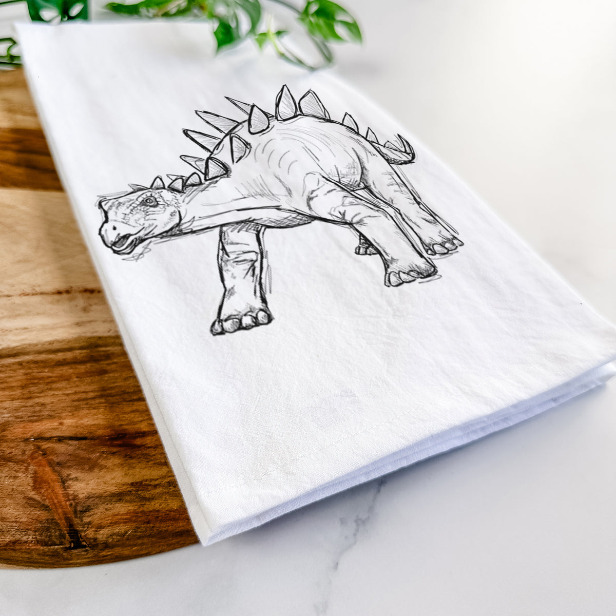 Close-up of a folded white tea towel on a wooden board, showing a black ink illustration of a Stegosaurus with plates along its back.