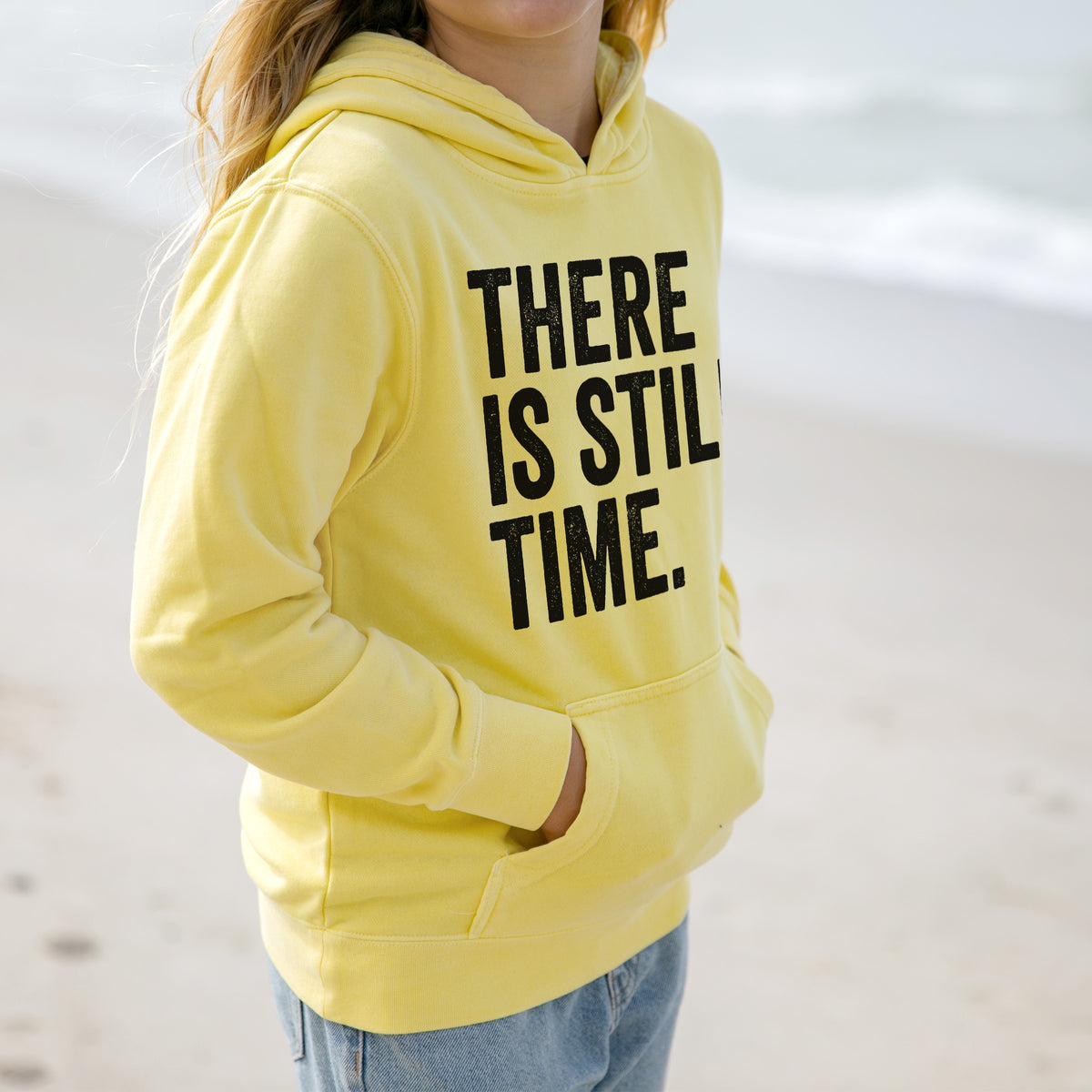 Girl wearing a youth pigment dyed hoodie printed with bold black words reading There Is Still Time.