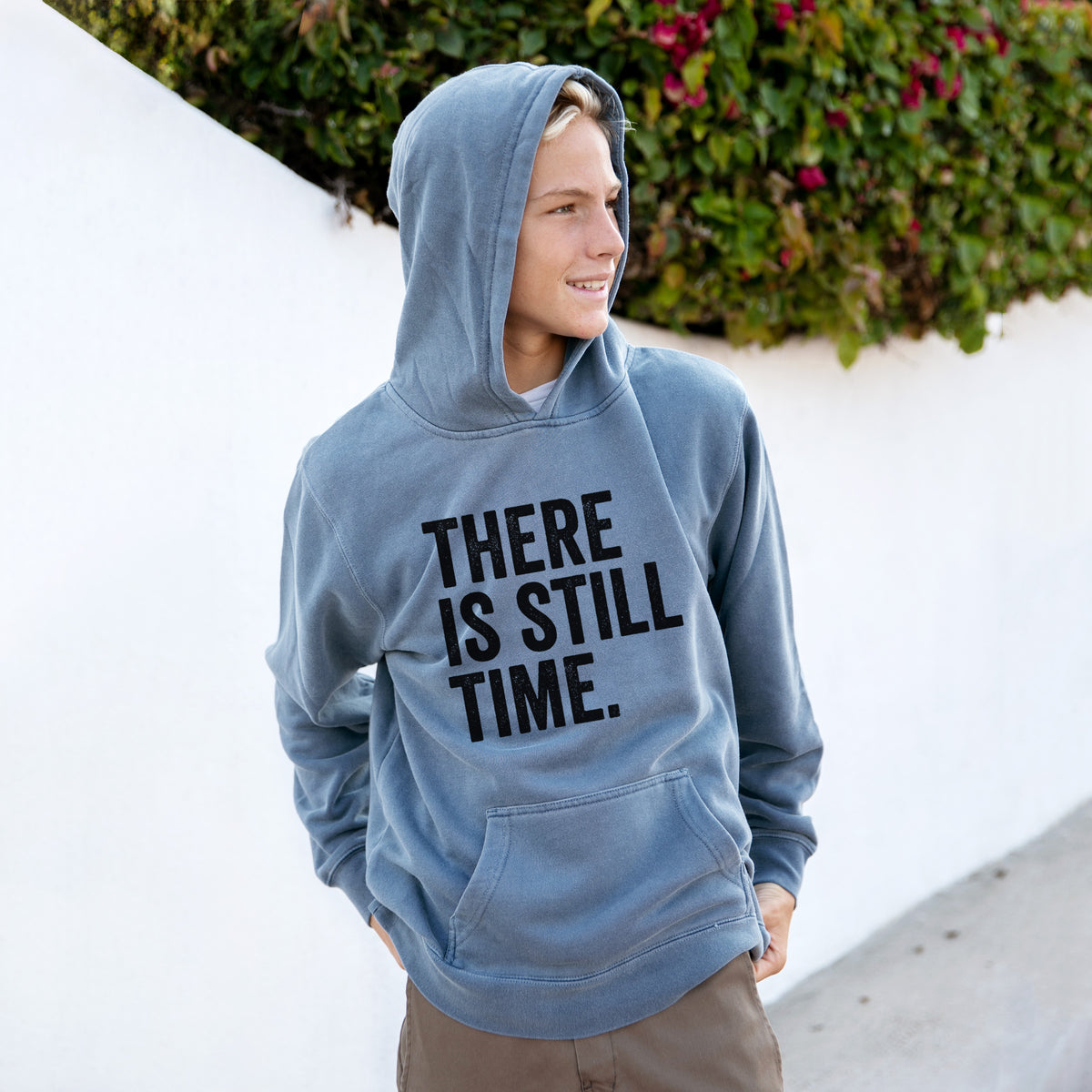 Boy wearing a youth pigment dyed hoodie printed with bold black words reading There Is Still Time.