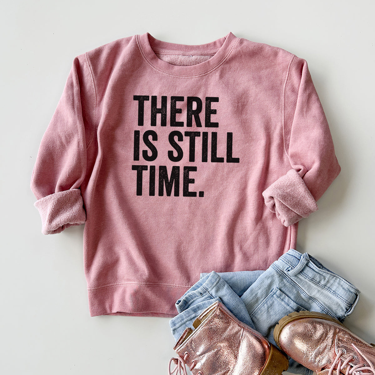 Youth crewneck sweatshirt printed with bold black words reading There Is Still Time, styled flat with jeans and boots.