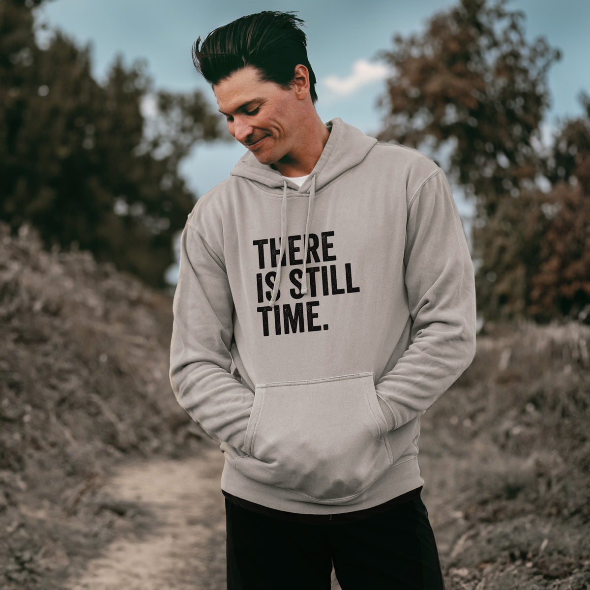 Man wearing a cement grey pigment dyed hoodie printed with bold black words reading There Is Still Time.