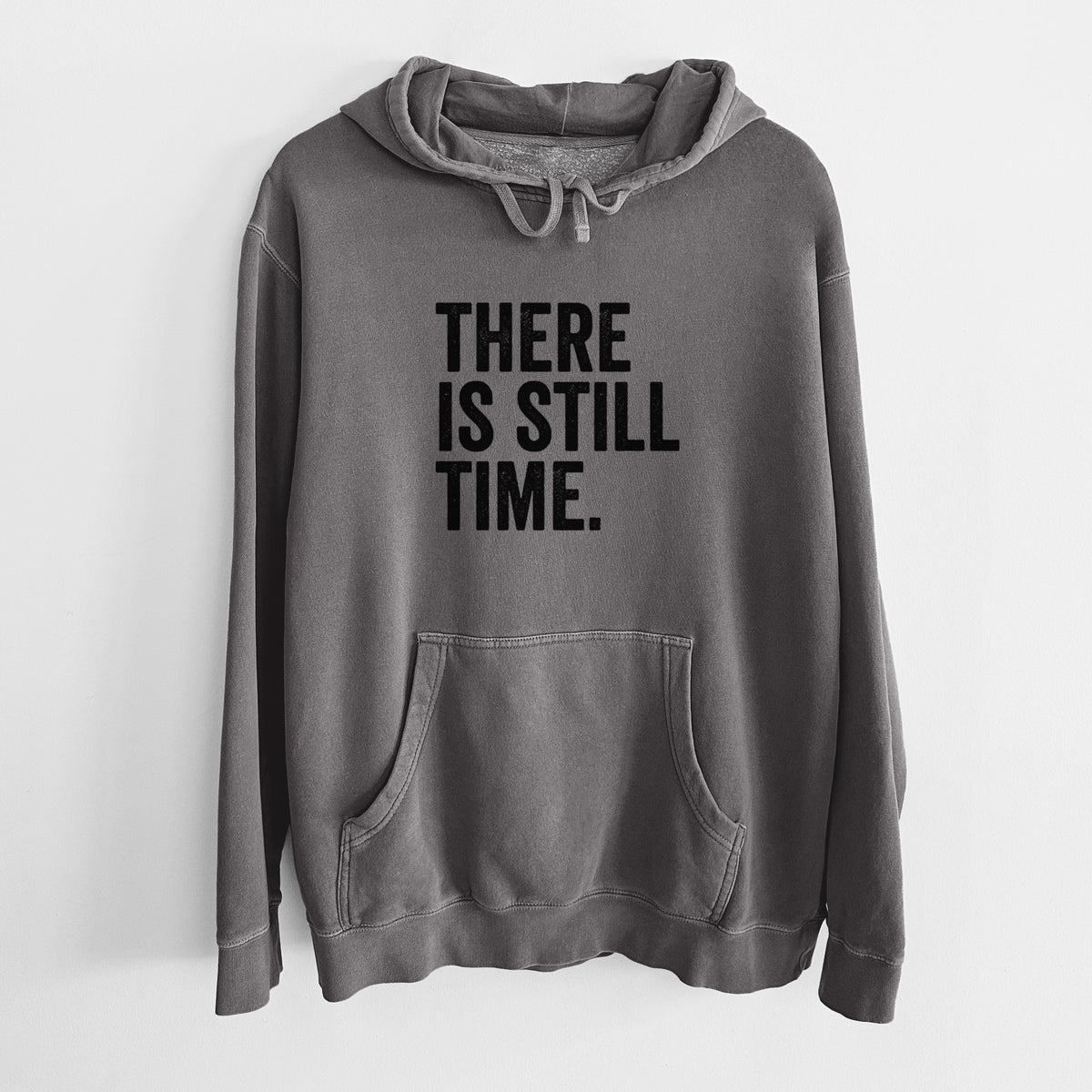 Faded black pigment dyed hoodie laid flat, printed with bold black words reading There Is Still Time.