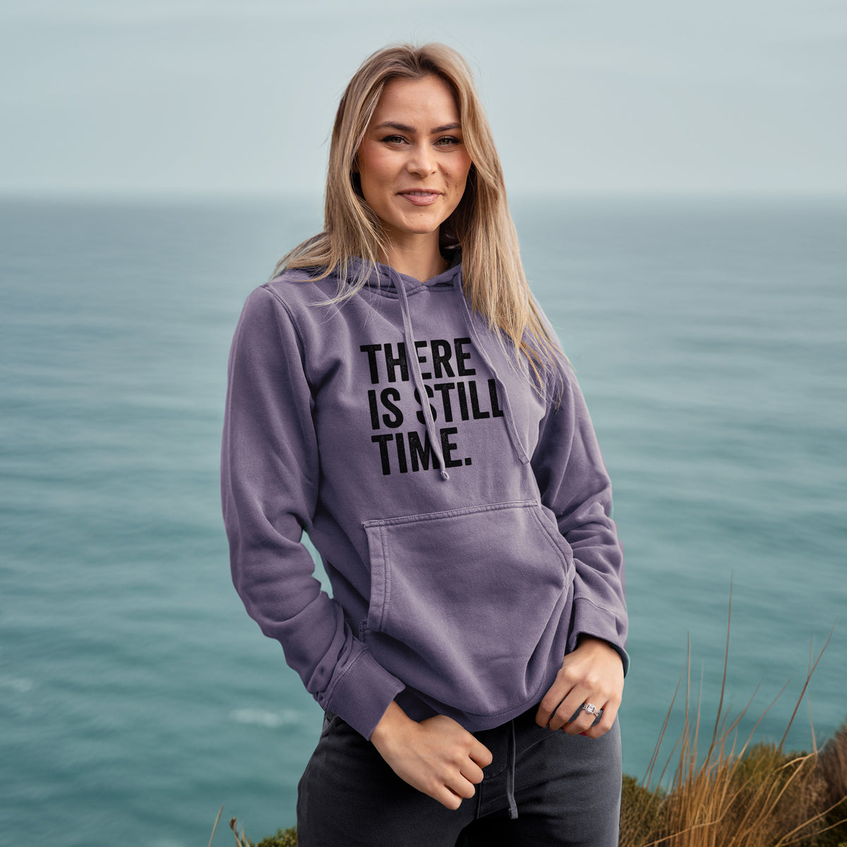 Woman wearing a plum purple pigment dyed hoodie printed with bold black words reading There Is Still Time.