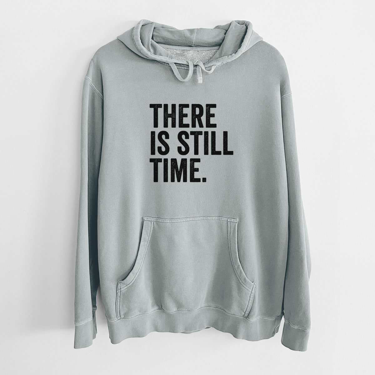 Sage green pigment dyed hoodie laid flat, printed with bold black words reading There Is Still Time.
