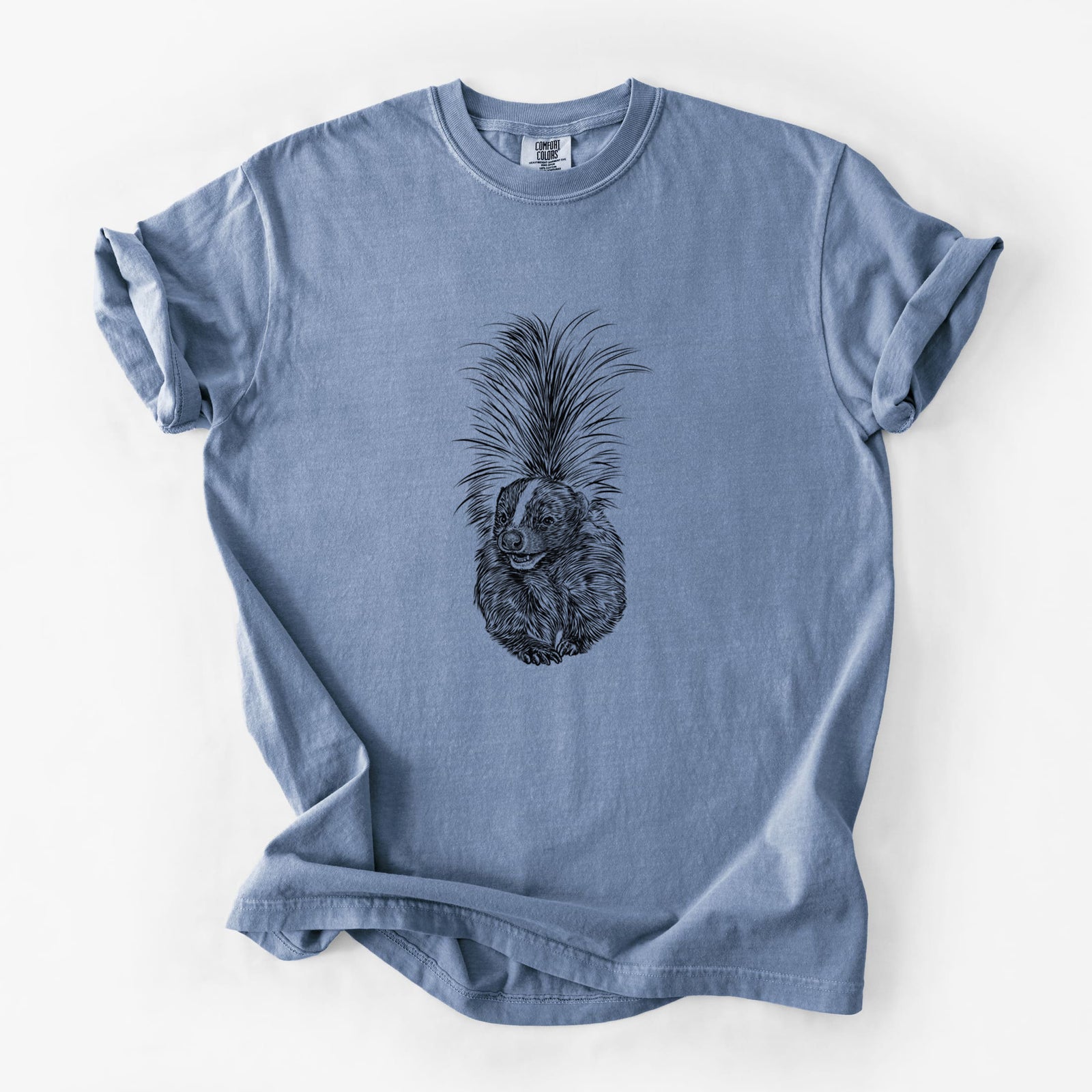 Blue jean Comfort Colors t-shirt laid flat, printed with a detailed black ink illustration of a striped skunk with its tail raised.