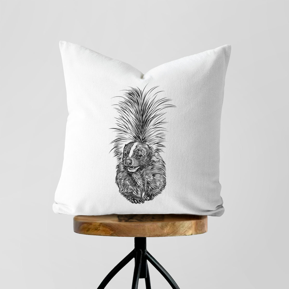 White pillow cover printed with a detailed black ink illustration of a striped skunk with its tail raised, on a wooden stool.