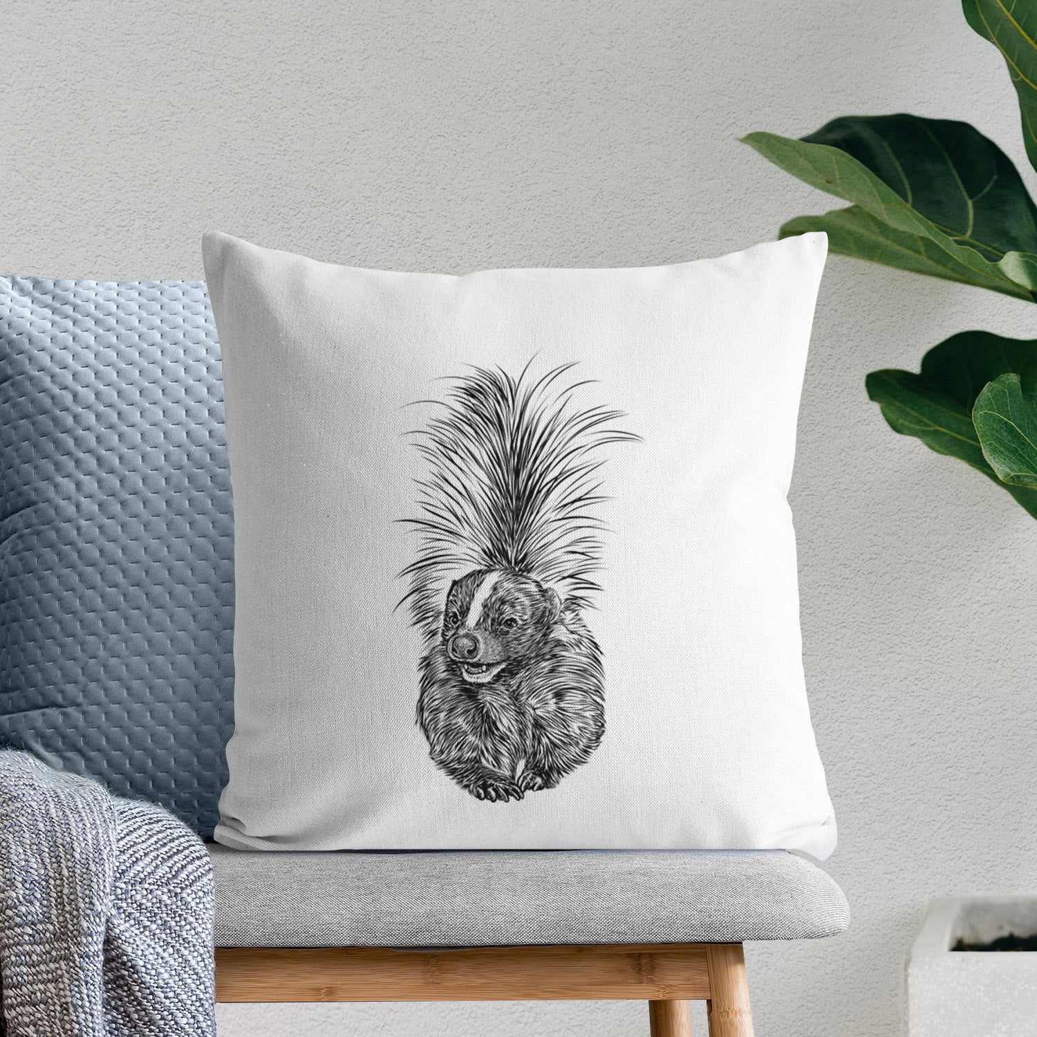 White pillow cover printed with a detailed black ink illustration of a striped skunk with its tail raised, on a bench beside plants.