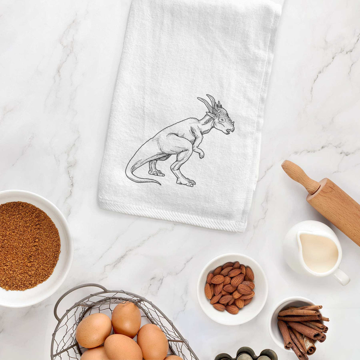 White hand towel printed with a black ink illustration of a Stygimoloch dinosaur with a spiky domed head, hanging on a cabinet door.
