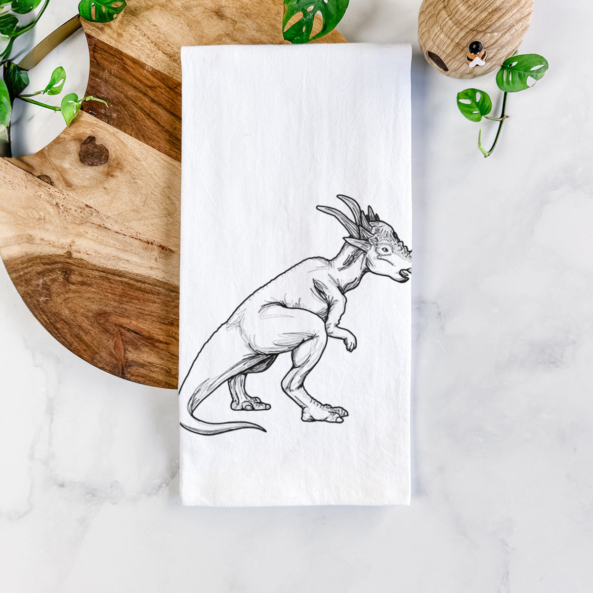 White cotton tea towel printed with a black ink illustration of a Stygimoloch dinosaur with a spiky domed head, on a marble counter beside a round wooden cutting board and greenery.