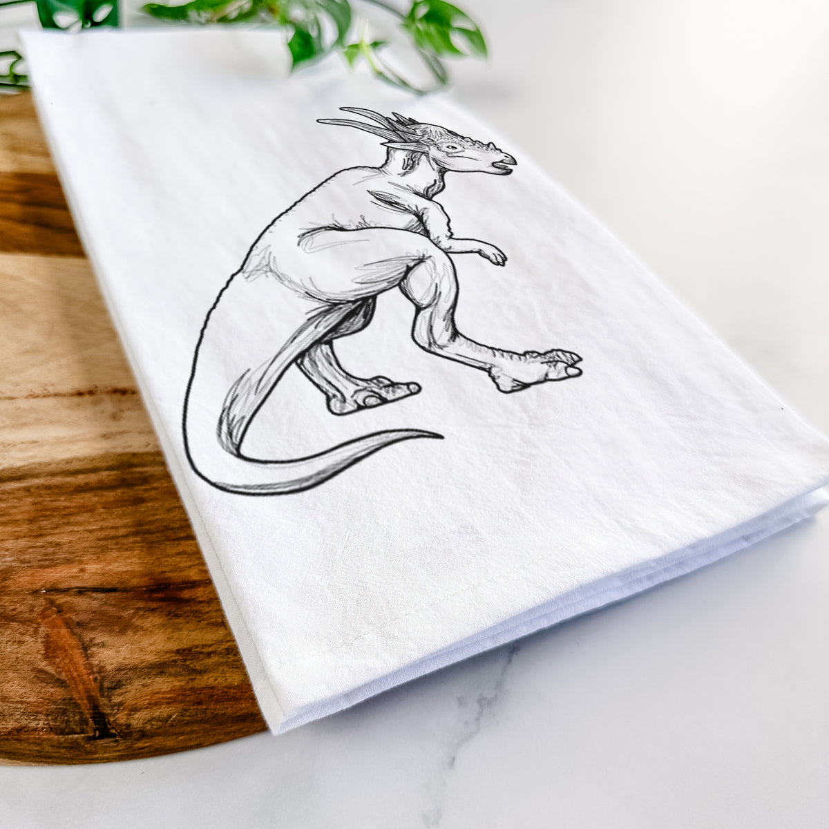 Close-up of a folded white tea towel on a wooden board, showing a black ink illustration of a Stygimoloch dinosaur with a spiky domed head.