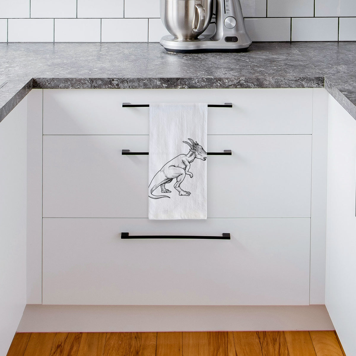 White tea towel printed with a black ink illustration of a Stygimoloch dinosaur with a spiky domed head, hanging from a drawer handle in a bright white kitchen.