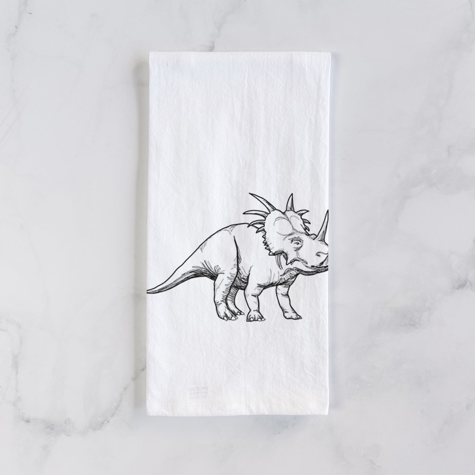 White cotton tea towel printed with a black ink illustration of a Styracosaurus with its spiked frill, on a marble counter beside a round wooden cutting board and greenery.