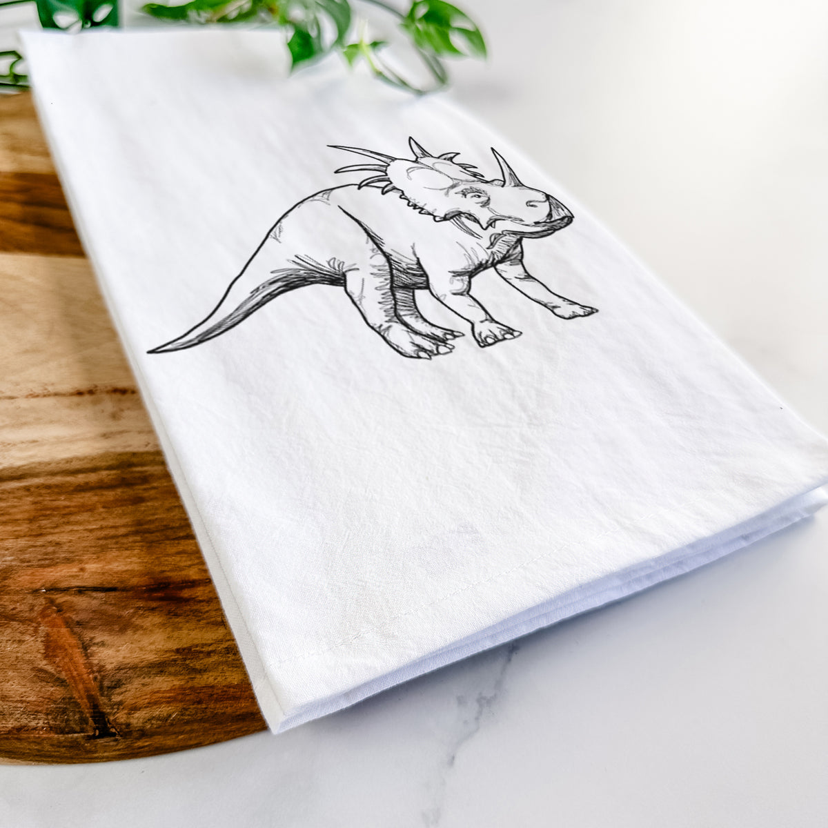 Close-up of a folded white tea towel on a wooden board, showing a black ink illustration of a Styracosaurus with its spiked frill.
