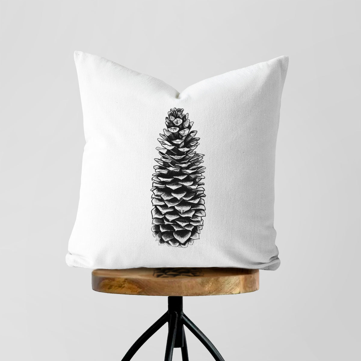 White pillow cover printed with a detailed black ink illustration of a long sugar pine cone, on a wooden stool.