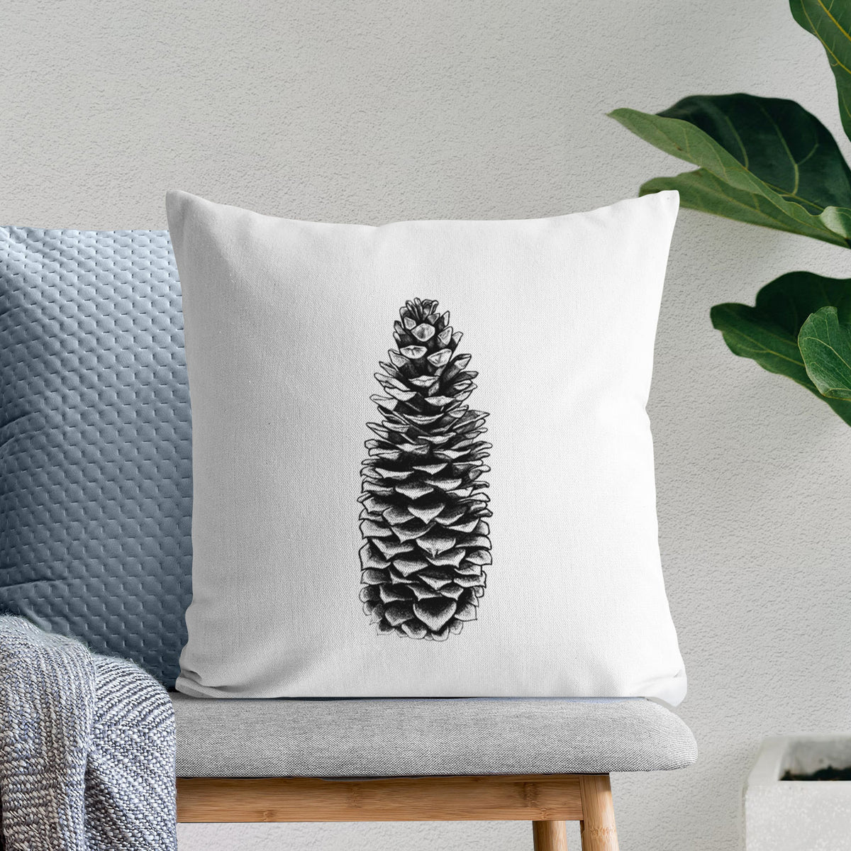 White pillow cover printed with a detailed black ink illustration of a long sugar pine cone, on a bench beside plants.