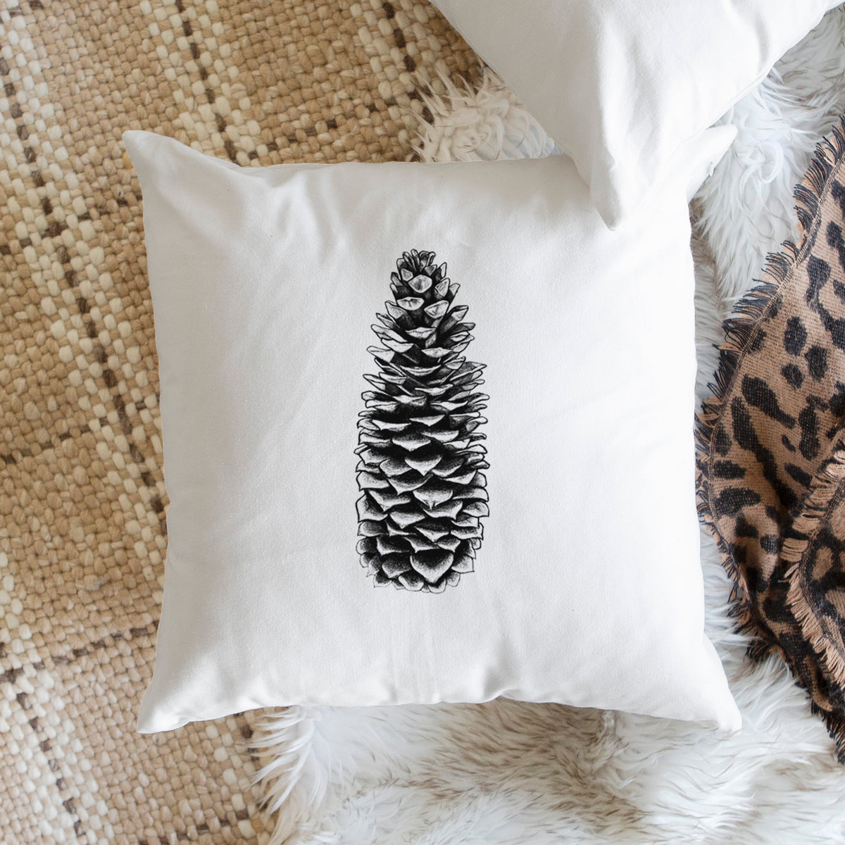 White pillow cover printed with a detailed black ink illustration of a long sugar pine cone, on a woven chair.