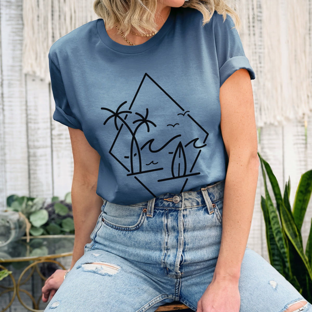 Woman wearing a steel blue lightweight cotton t-shirt printed with black line art of palm trees, ocean waves, birds and a surfboard inside a diamond outline.