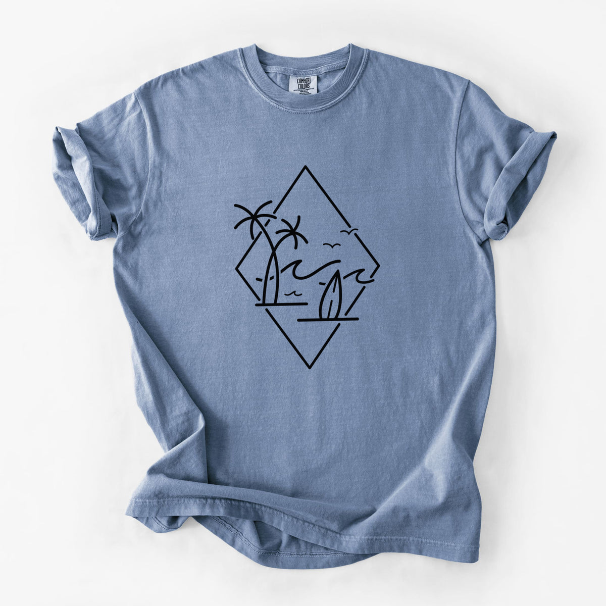 Blue jean Comfort Colors t-shirt laid flat, printed with black line art of palm trees, ocean waves, birds and a surfboard inside a diamond outline.