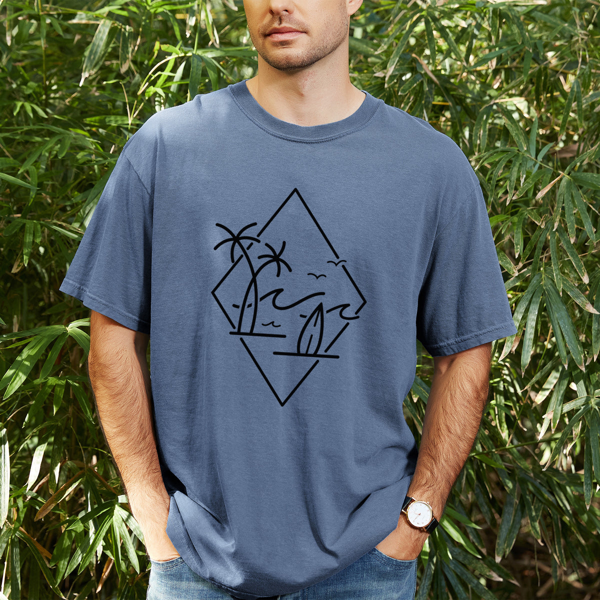 Man wearing a Comfort Colors t-shirt printed with black line art of palm trees, ocean waves, birds and a surfboard inside a diamond outline, outdoors.