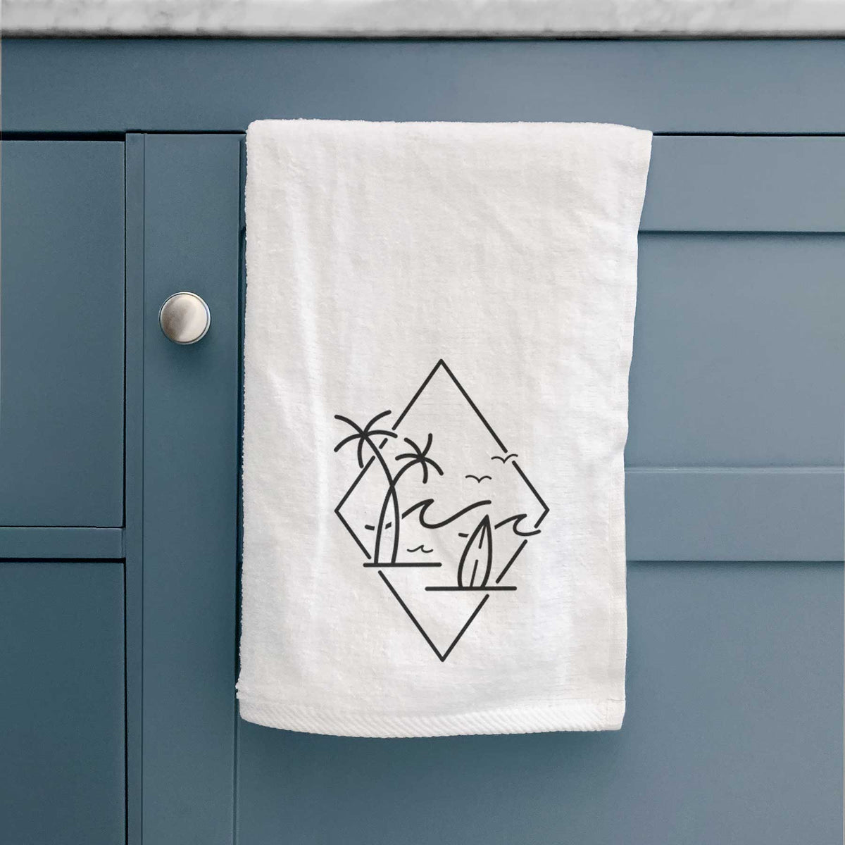 White hand towel printed with black line art of palm trees, ocean waves, birds and a surfboard inside a diamond outline, hanging on a cabinet door.