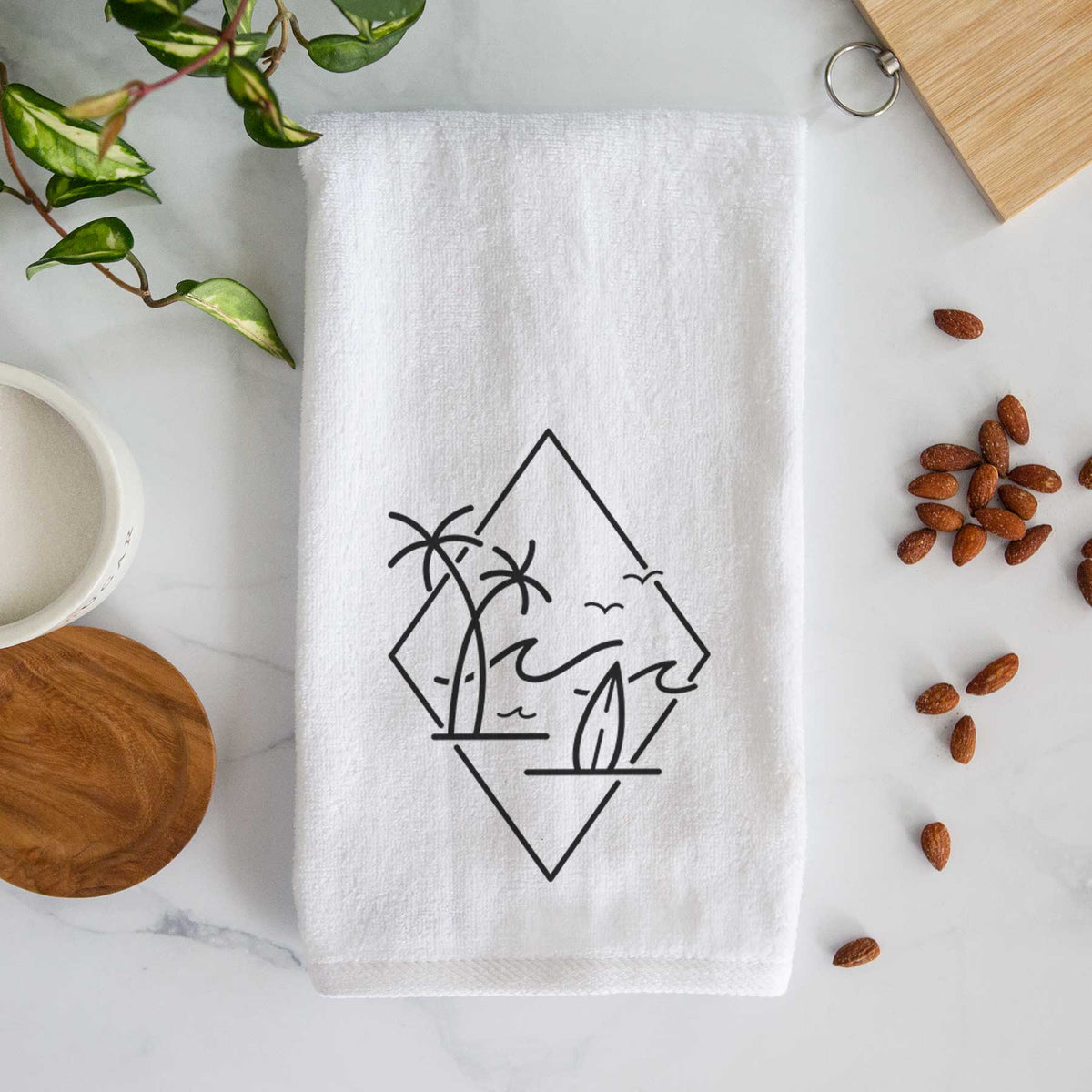 White hand towel printed with black line art of palm trees, ocean waves, birds and a surfboard inside a diamond outline.
