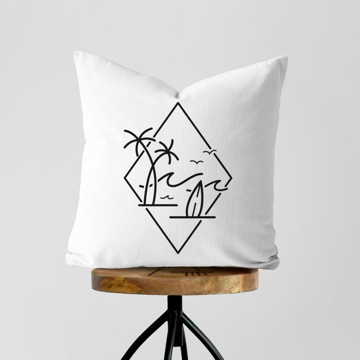 White pillow cover printed with black line art of palm trees, ocean waves, birds and a surfboard inside a diamond outline, on a wooden stool.
